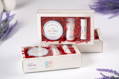 Two Lavender Gift Sets, each with Lavender Body Butter and Lip Balm, are displayed in clear boxes with red shredded paper on a white surface, accented by lavender flowers in the background.