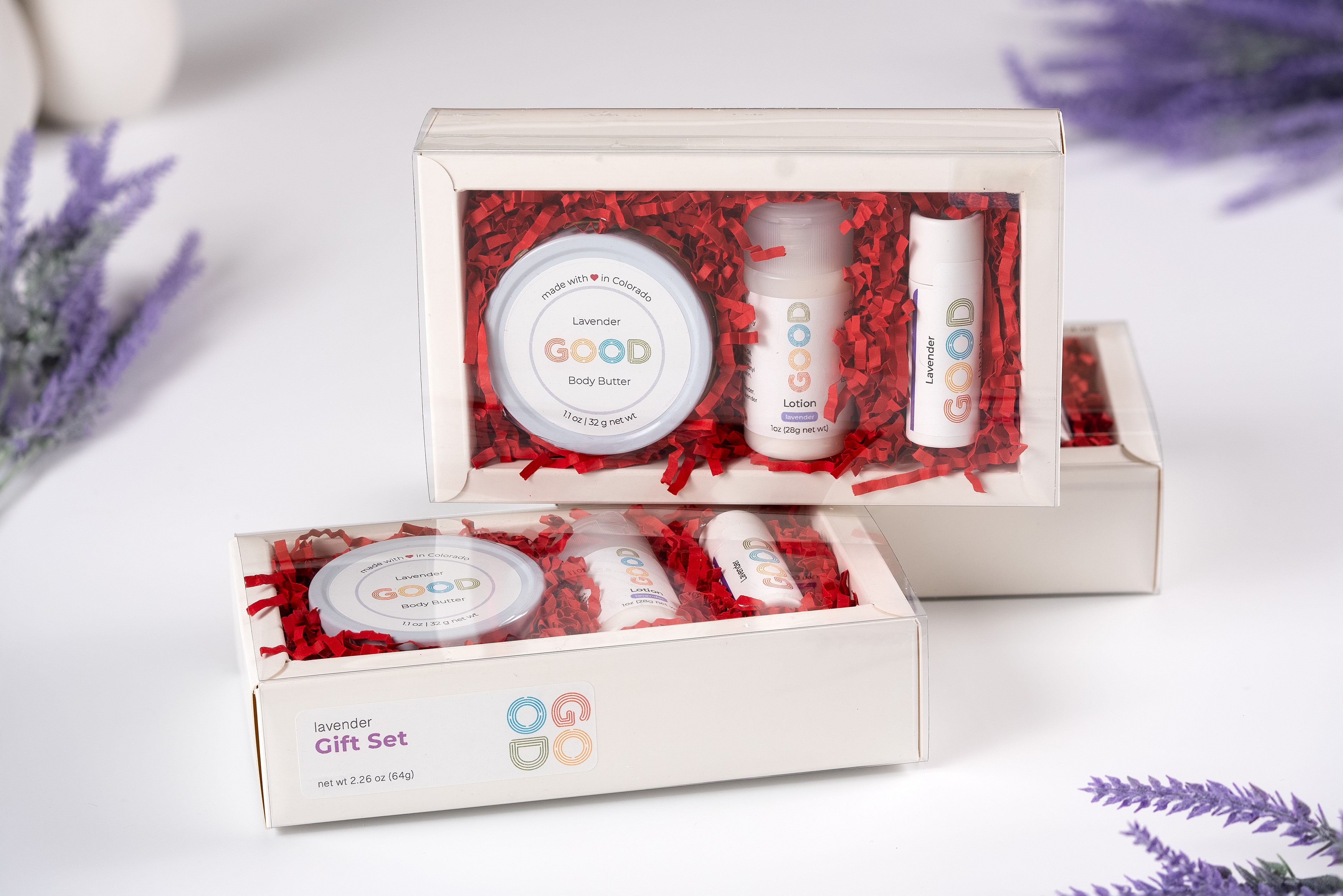 Two Lavender Gift Sets, each with Lavender Body Butter and Lip Balm, are displayed in clear boxes with red shredded paper on a white surface, accented by lavender flowers in the background.