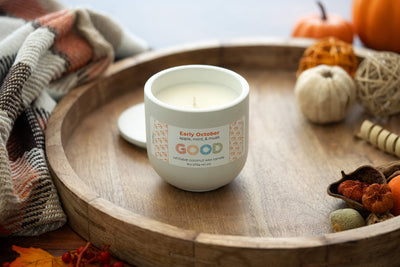 The Early October candle—a white, refillable coconut wax candle in a glass jar—features notes of apple, mint, and musk. It's shown on a wood tray with fall accents: pumpkins, dried leaves, and a plaid blanket for a cozy autumn vibe.