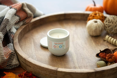 The Early October candle, made from natural coconut wax in a white concrete jar, rests on a wooden tray among autumn décor such as small pumpkins, dried leaves, and a plaid blanket.