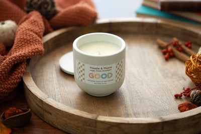 A Thistle & Thorn white coconut wax candle labeled GOOD sits on a round wooden tray, surrounded by autumn decor: an orange knit blanket, berries, cinnamon sticks, and books.