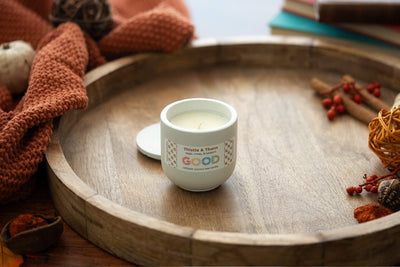 A small white Thistle & Thorn coconut wax candle in a jar rests on a round wooden tray, surrounded by autumn decor: an orange knit cloth, berries, cinnamon sticks, and stacked books for a cozy ambiance.
