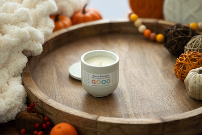 The Berry Booquet coconut wax candle in a concrete jar rests on a wooden tray with small pumpkins, decorative balls, a cozy white blanket, and a colorful bead garland, filling the space with phthalate-free autumn fragrances.