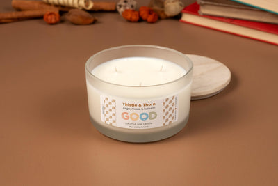 A white, three-wick coconut wax candle labeled Thistle & Thorn sits in a frosted glass jar with a wooden lid nearby. The refillable candle rests on a brown surface, surrounded by books and dried botanicals.
