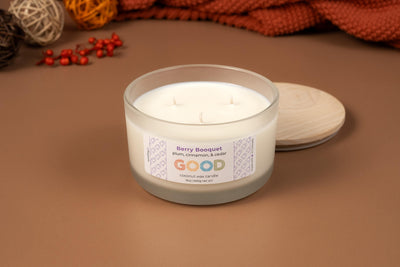 A white three-wick coconut wax candle labeled Berry Booquet sits in a frosted glass jar on a brown surface, its wooden lid beside it and decorative items in the background.