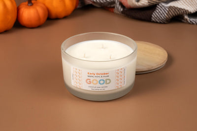 The white three-wick Early October candle, made from natural coconut wax and scented with apple, mint, and musk, sits on a brown surface beside its wooden lid, surrounded by pumpkins and a plaid blanket in the background.