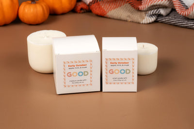 Two white Candle Refills boxes labeled “Early October” rest on a brown surface with white candles, small pumpkins, and a plaid blanket. These coconut-wax refills feature apple, mint & musk scents in biodegradable packaging.