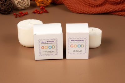 Two white Candle Refills boxes labeled Berry Bouquet with "GOOD" in colorful letters, and two white candles in concrete vessels, sit on a brown surface with decorative balls and fabric in the background.
