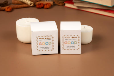 Two boxes labeled “Thistle & Thorn: sage, moss, & balsam” Candle Refills with “GOOD” below sit on a brown surface in biodegradable packaging. White candles, cinnamon sticks, and books appear in the background.