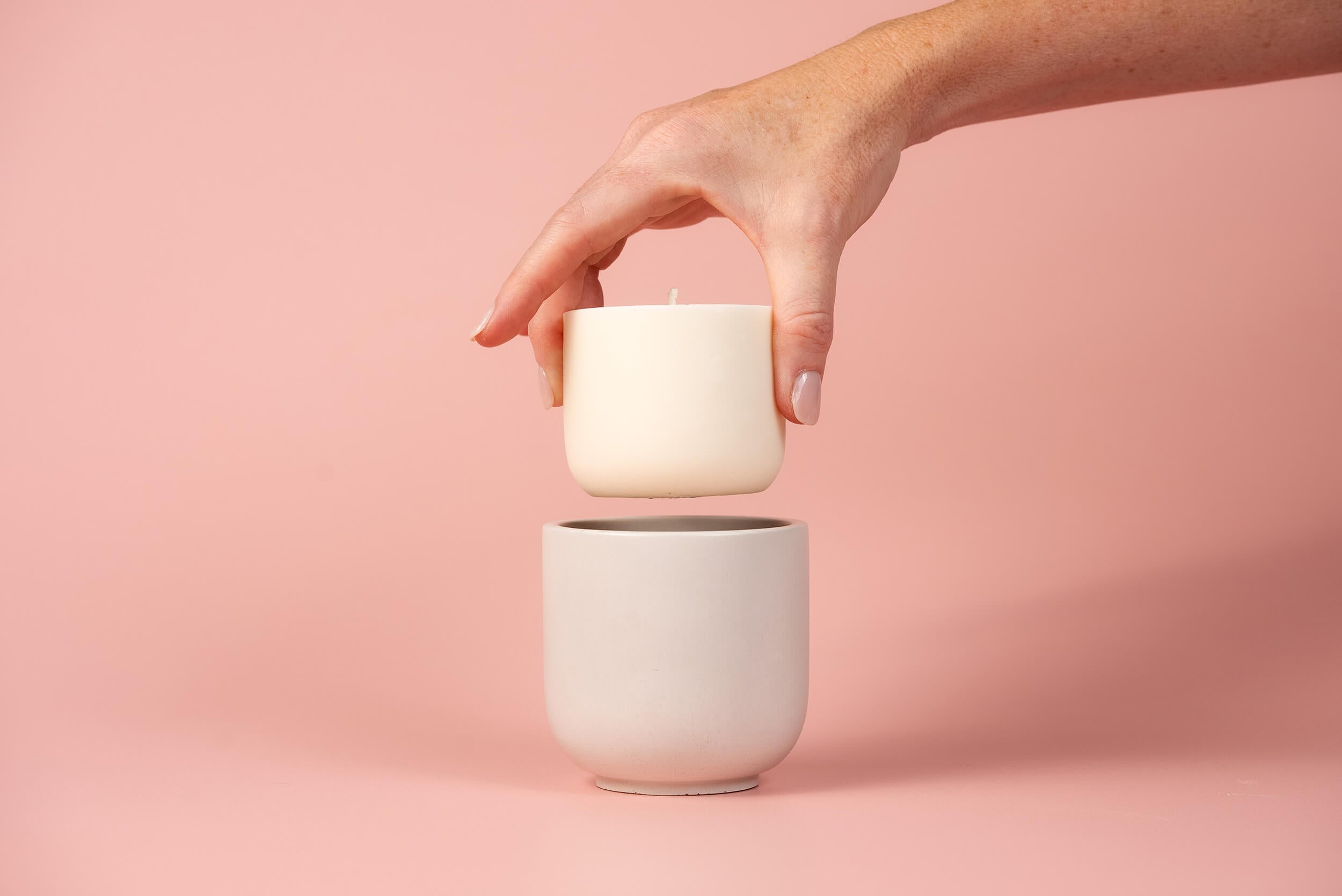 A hand holding a small white candle above a light gray ceramic cup against a pink background.