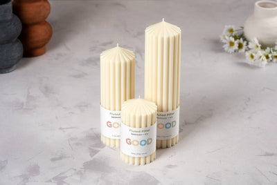 Three cream-colored Fluted Pillar candles of different heights stand on a light gray surface, each with a "GOOD" label. Handmade, they are set with white daisies and pottery pieces in the background.