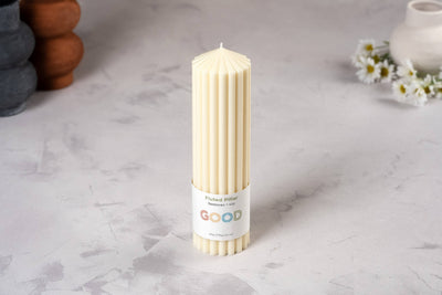 A tall, cream-colored Fluted Pillar Candle made from beeswax and soy stands upright on a light gray surface. The label reads "GOOD." Daisies and ceramic objects are softly blurred in the background.