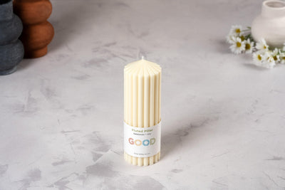 A cream-colored, fluted beeswax pillar candle from our Fluted Pillar Candles set stands on a light gray surface. Its label reads “Fluted pillar, beeswax, 15H, GOOD.” Daisies and ceramic vases blur in the background.