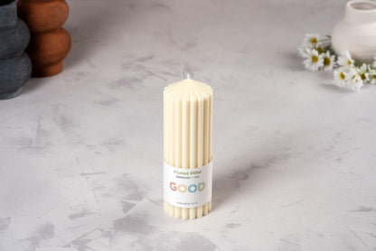 A cream-colored, fluted beeswax pillar candle from our Fluted Pillar Candles set stands on a light gray surface. Its label reads “Fluted pillar, beeswax, 15H, GOOD.” Daisies and ceramic vases blur in the background.