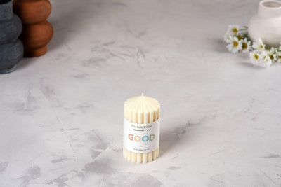 A cream fluted candle from our Fluted Pillar Candles set labeled "GOOD" rests on a light, textured surface, with white daisies and ceramic vessels in the background enhancing the charm of these elegant unscented candles.
