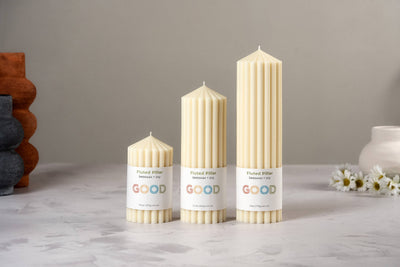 Three Fluted Pillar Candles in ivory beeswax, each a different height, are arranged on a light surface. Each candle features a "Fluted Pillar GOOD" label; decorative vases and white flowers appear in the background.