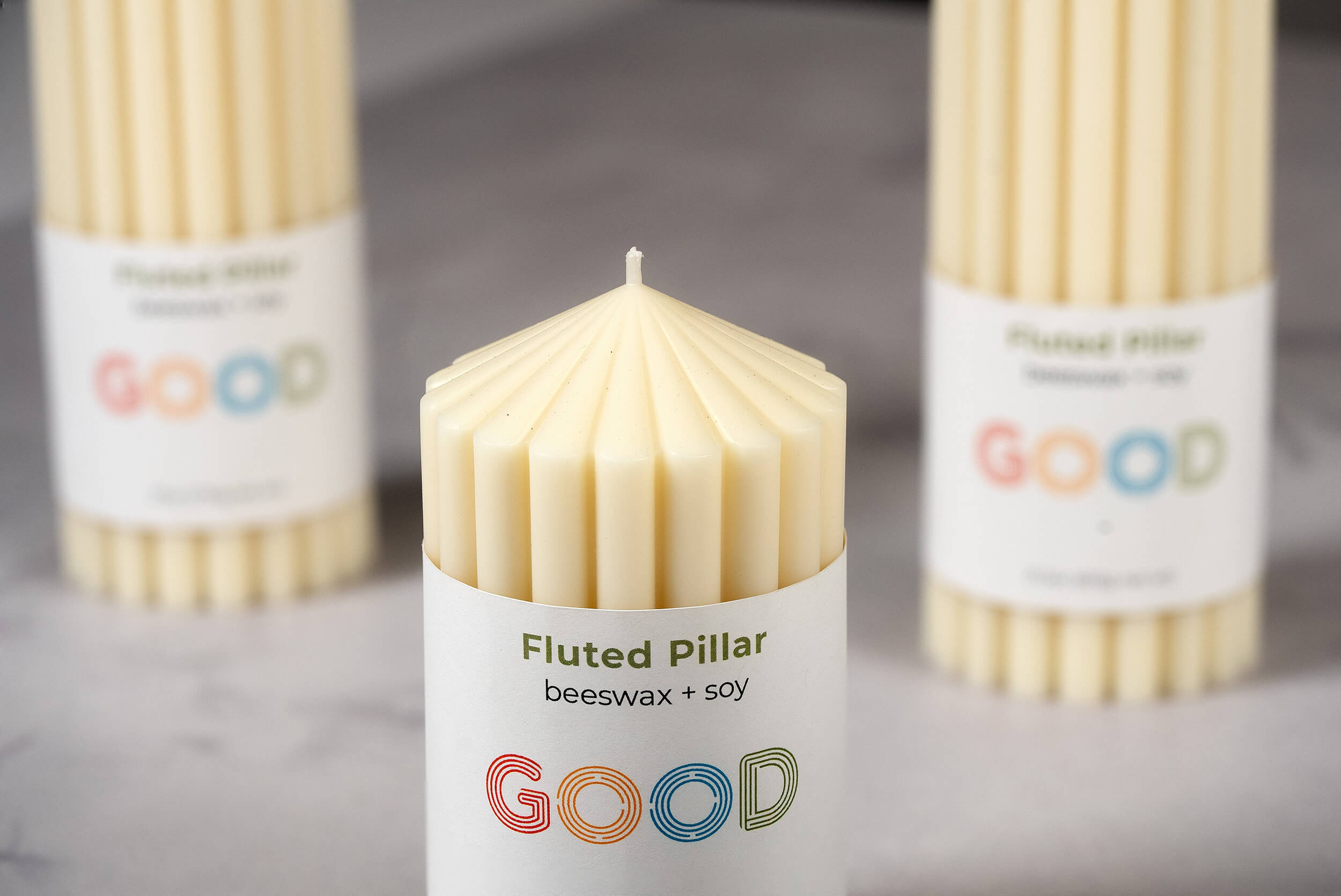 A close-up of a fluted pillar candle made from beeswax and soy, labeled "GOOD," with two unscented Fluted Pillar Candles softly blurred in the background on a light surface.