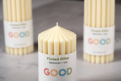 A close-up of a fluted pillar candle made from beeswax and soy, labeled "GOOD," with two unscented Fluted Pillar Candles softly blurred in the background on a light surface.