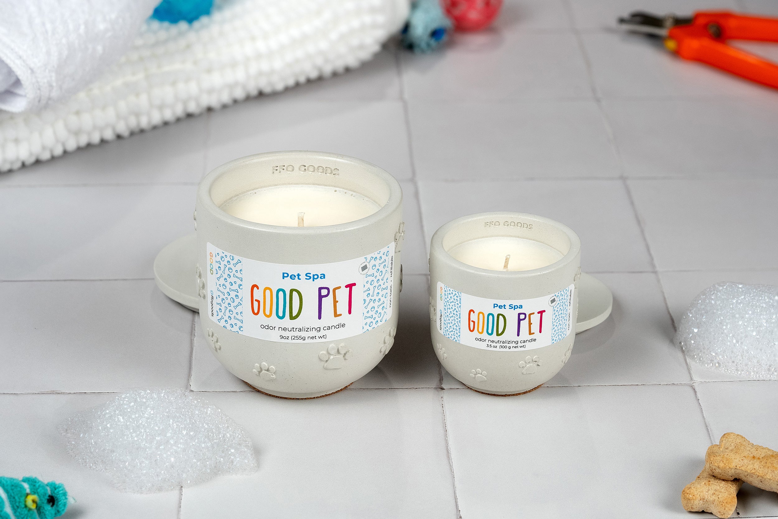 Two white Pet Spa candles in GOOD PET mugs rest on tiles with soap bubbles, a dog biscuit, pet toys, and grooming scissors nearby, creating a spa setting with odor-eliminating scents for your furry companion.
