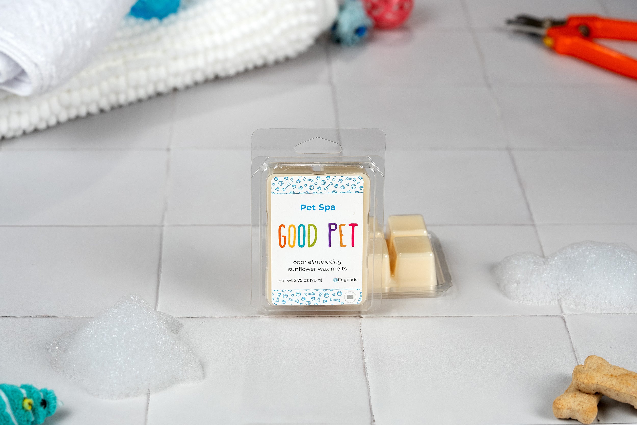 A package of Pet Spa Wax Melts is displayed on tiles alongside soap suds, a towel, dog treats, and grooming scissors, emphasizing a cozy pet spa atmosphere with plant-based, odor-eliminating scents.