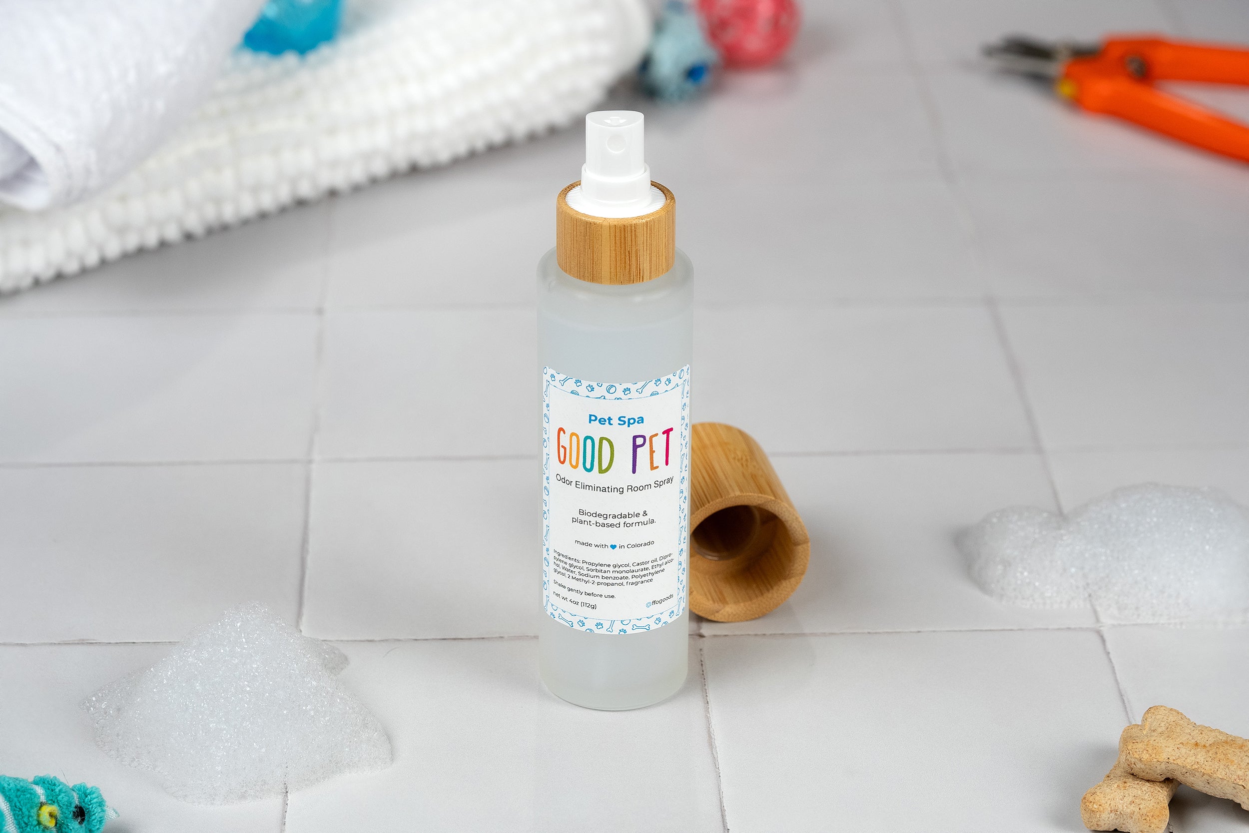 A bottle of Good Pet Room Spray, a plant-based air freshener, sits uncapped on a tiled surface. Surrounding it are soap bubbles, a towel, pet treats, grooming tools, and a blue toy—creating a cozy pet spa atmosphere.