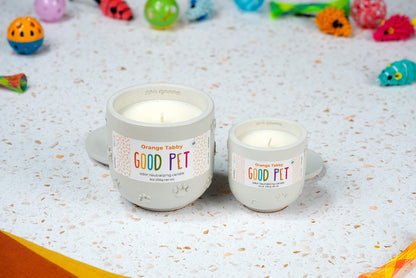 Two hand-poured Orange Tabby candles in light gray containers with paw prints sit on a white speckled surface, surrounded by colorful cat toys. The left candle is larger; both feature a crisp white color.