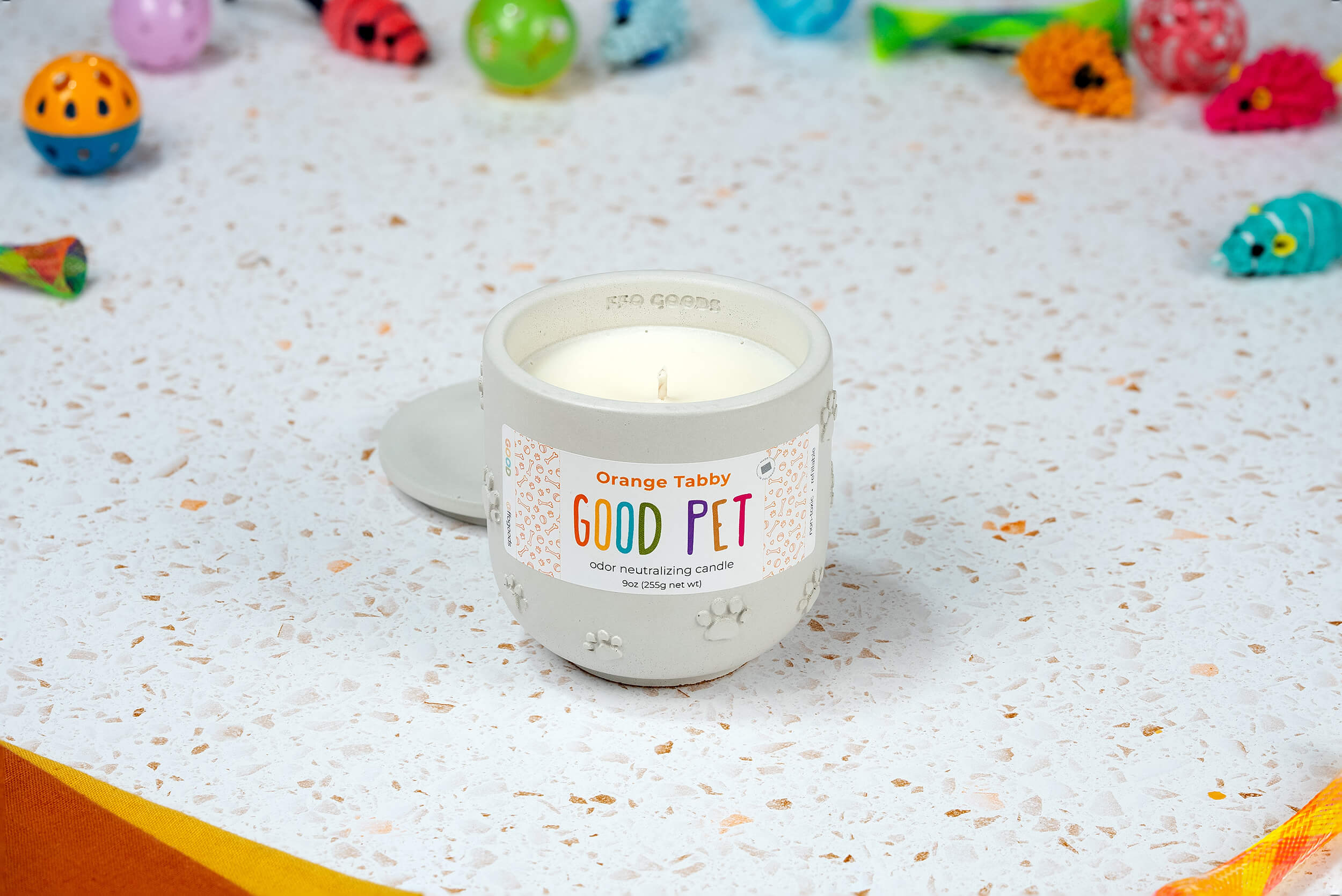 A hand-poured white candle called Orange Tabby, in a ceramic container labeled GOOD PET odor neutralizing candle, sits on a speckled surface with colorful pet toys in the background.