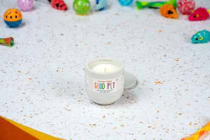 An Orange Tabby Good Pet odor-eliminating candle in a small mug rests on a terrazzo surface, with colorful cat toys in the background.