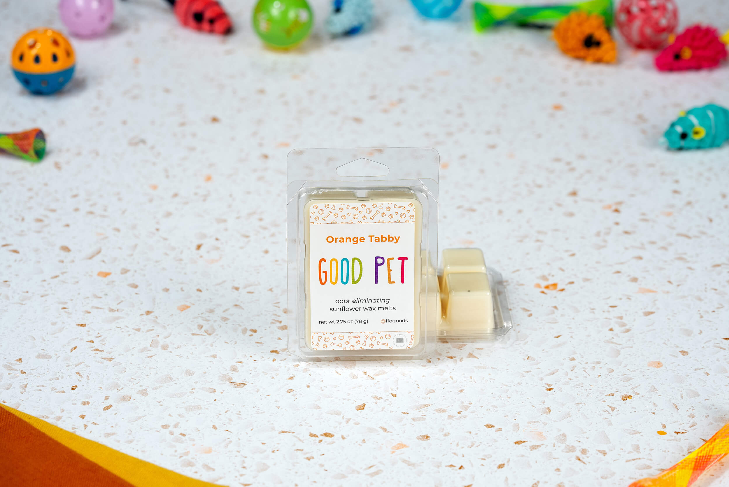 Orange Tabby Wax Melts sit on a speckled surface with colorful cat toys. These plant-based wax melts eliminate odors and fill your space with fresh, pet-friendly scents for a delightful home environment.