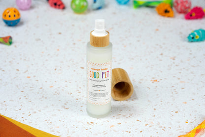 A Good Pet Room Spray with a bamboo cap sits on a terrazzo surface. The cap is off beside the bottle, colorful pet toys and fabric behind. This plant-based air freshener brightens any pet space.