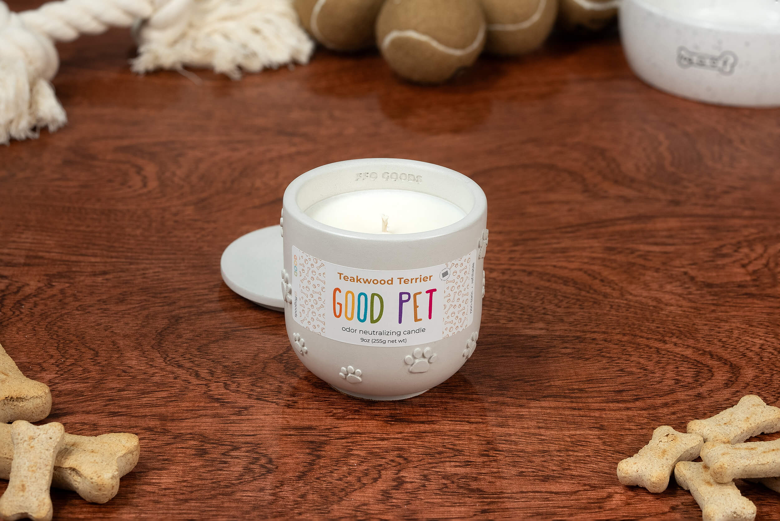 The Teakwood Terrier candle sits on a wooden surface among dog toys, a white bowl, and bone-shaped treats. Its odor-eliminating scent and nearby lid make any pet area fresh and inviting.