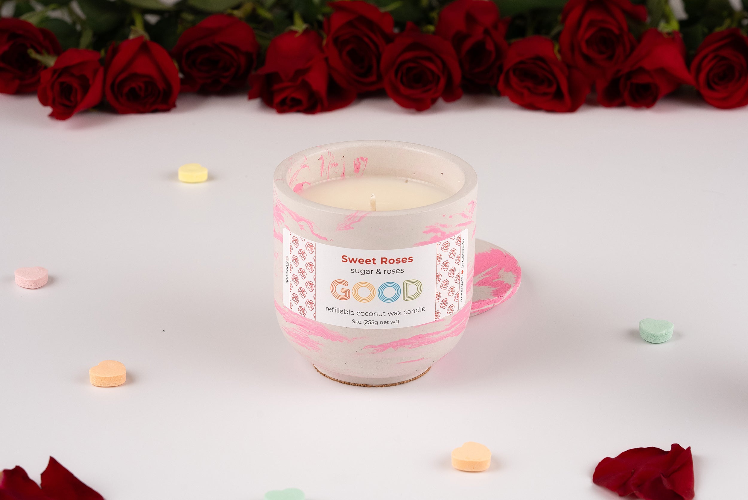 The Sweet Roses candle features a rose scent and comes in a white, pink-patterned glass jar. Displayed on a white surface with pastel candies, it’s set before a row of red roses.