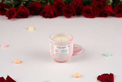 The Sweet Roses candle, featuring a white rose scent and pink marbled holder, rests on a white surface surrounded by heart candies and red rose petals, with red roses arranged in the background.