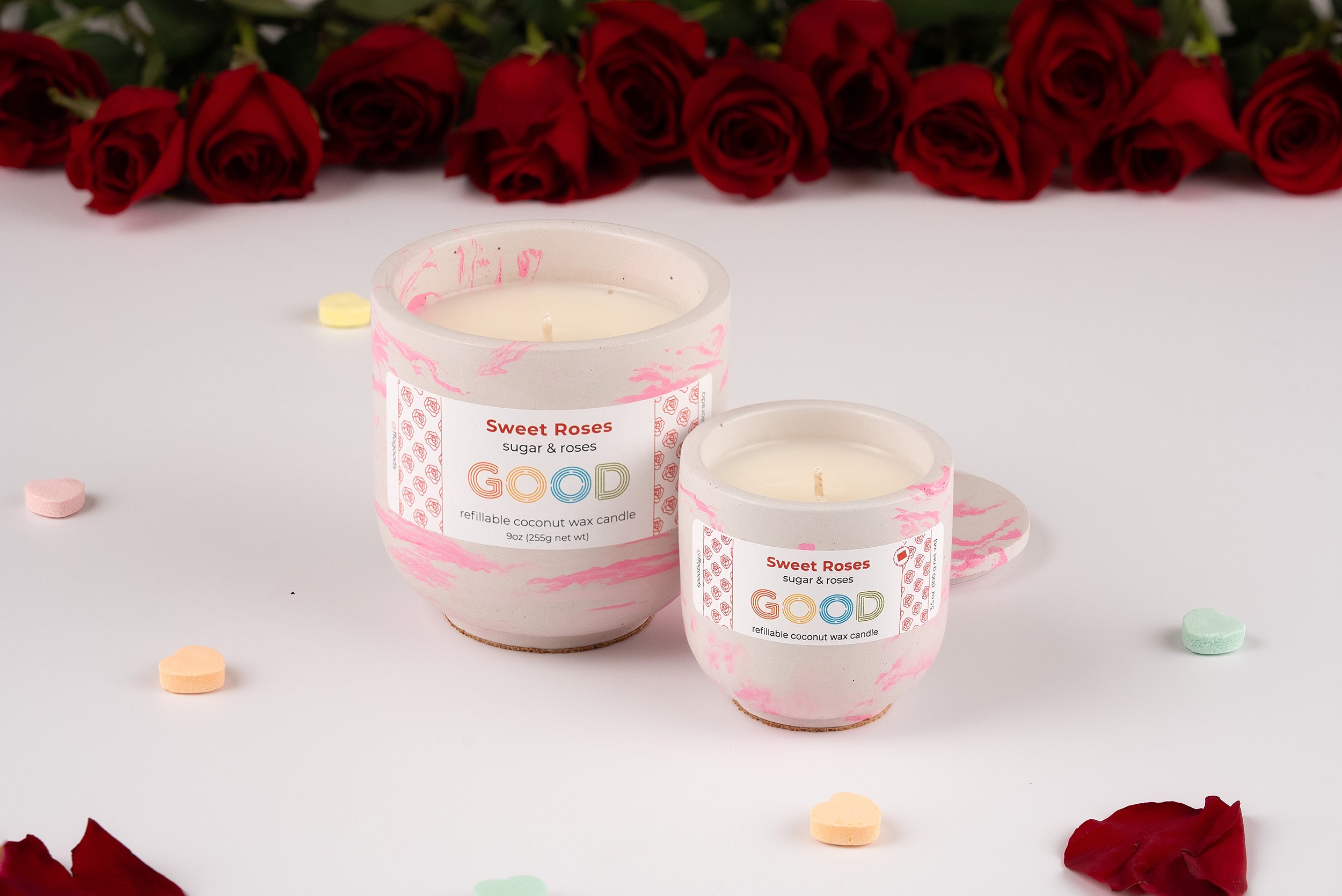 Two white handmade Sweet Roses candles rest on a white surface with red roses in the background and colorful heart-shaped candies scattered around, enhancing their beautiful rose scent.