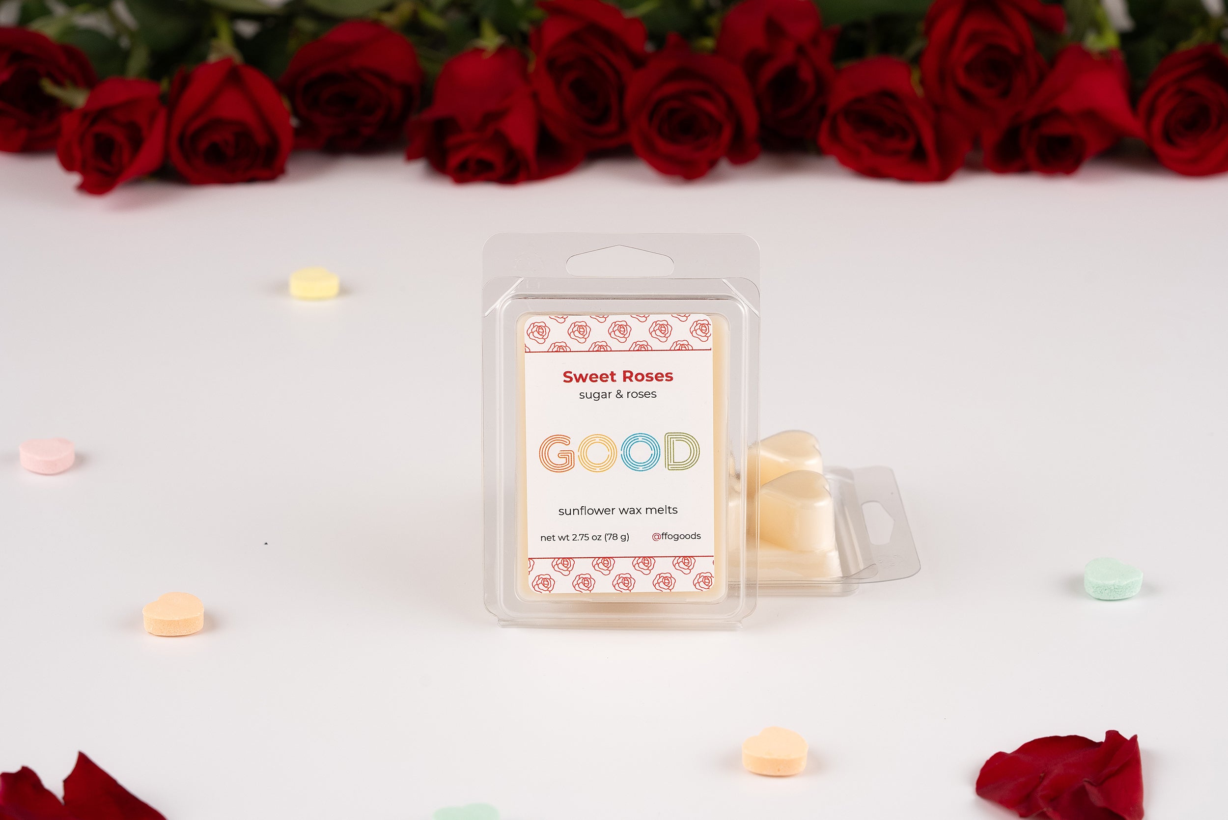 A package of Sweet Roses Wax Melts stands upright on a white surface, surrounded by heart-shaped candies, rose petals, and a row of red roses in the background.