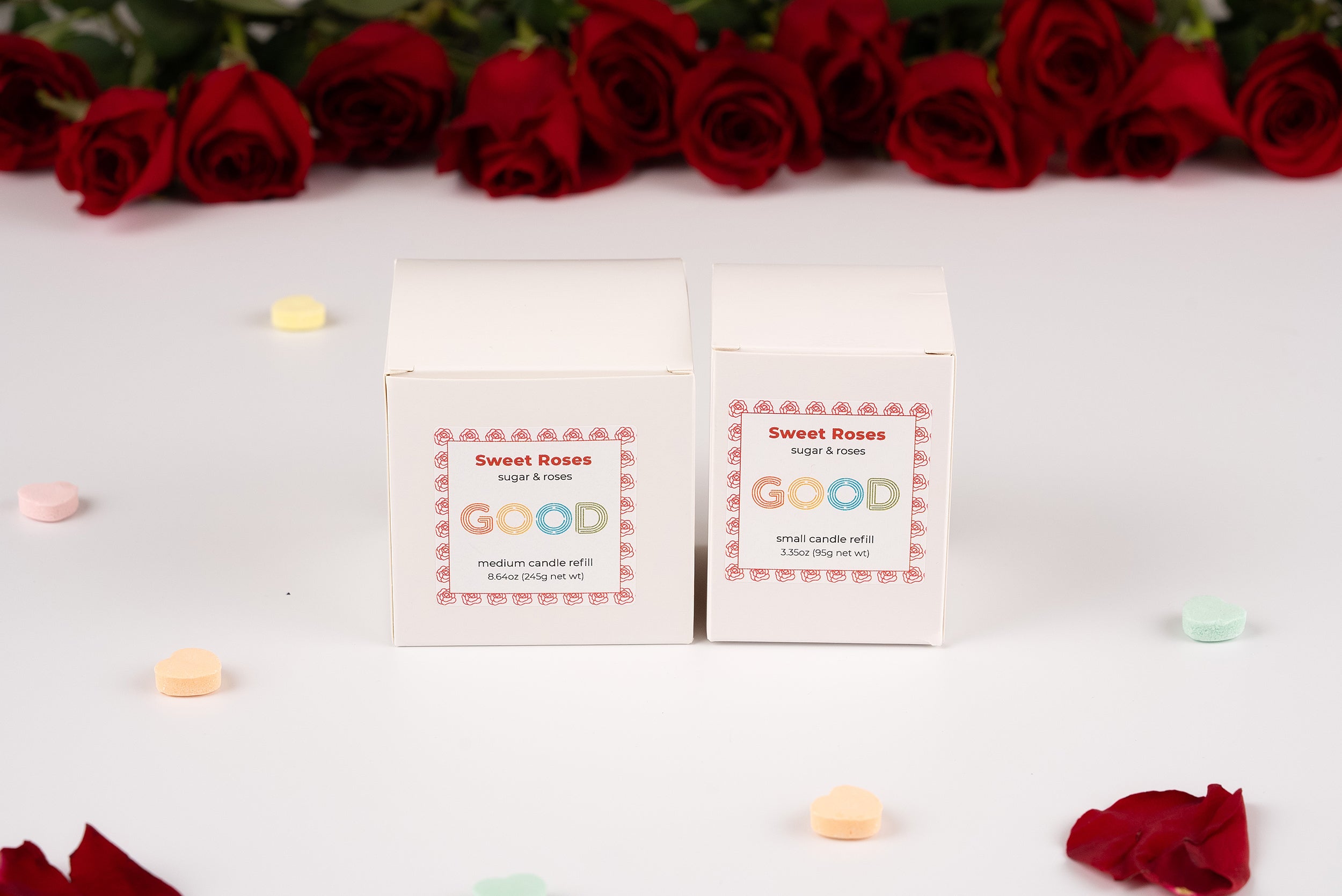 Two white boxes labeled Candle Refills sit on a white surface with pastel candy hearts and rose petals. Concrete candle vessels complete the scene, while a row of red roses is arranged in the background.