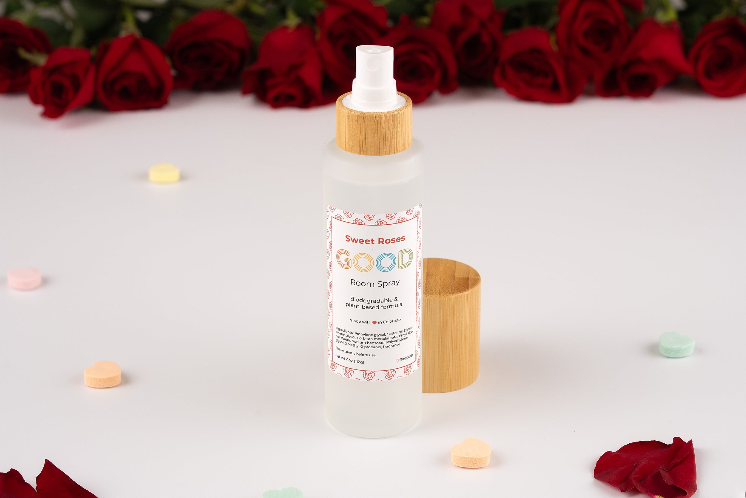 A bottle of Room Spray with a biodegradable, rose-scented formula and bamboo cap sits on a white surface, surrounded by red roses and pastel heart-shaped candies. The label highlights its Biodegradable & Rose Scented Formula.