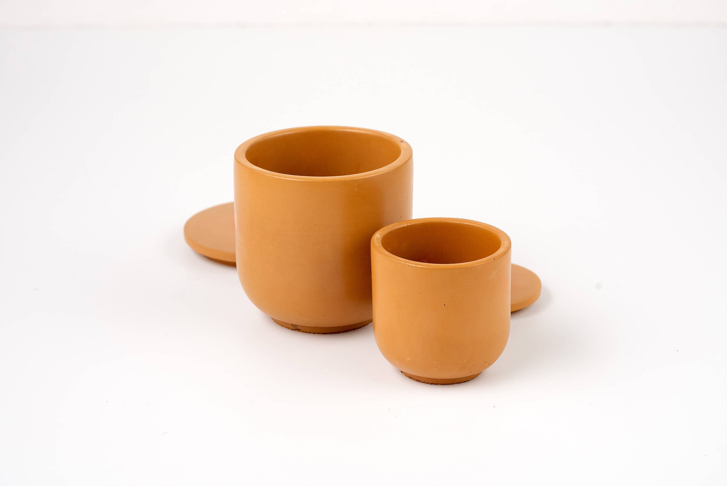 Two Wheat Buff Candle Vessels in brown ceramic, each a different size with matching round lids, are arranged on a white surface. The smaller vessel is positioned in front of the larger one.