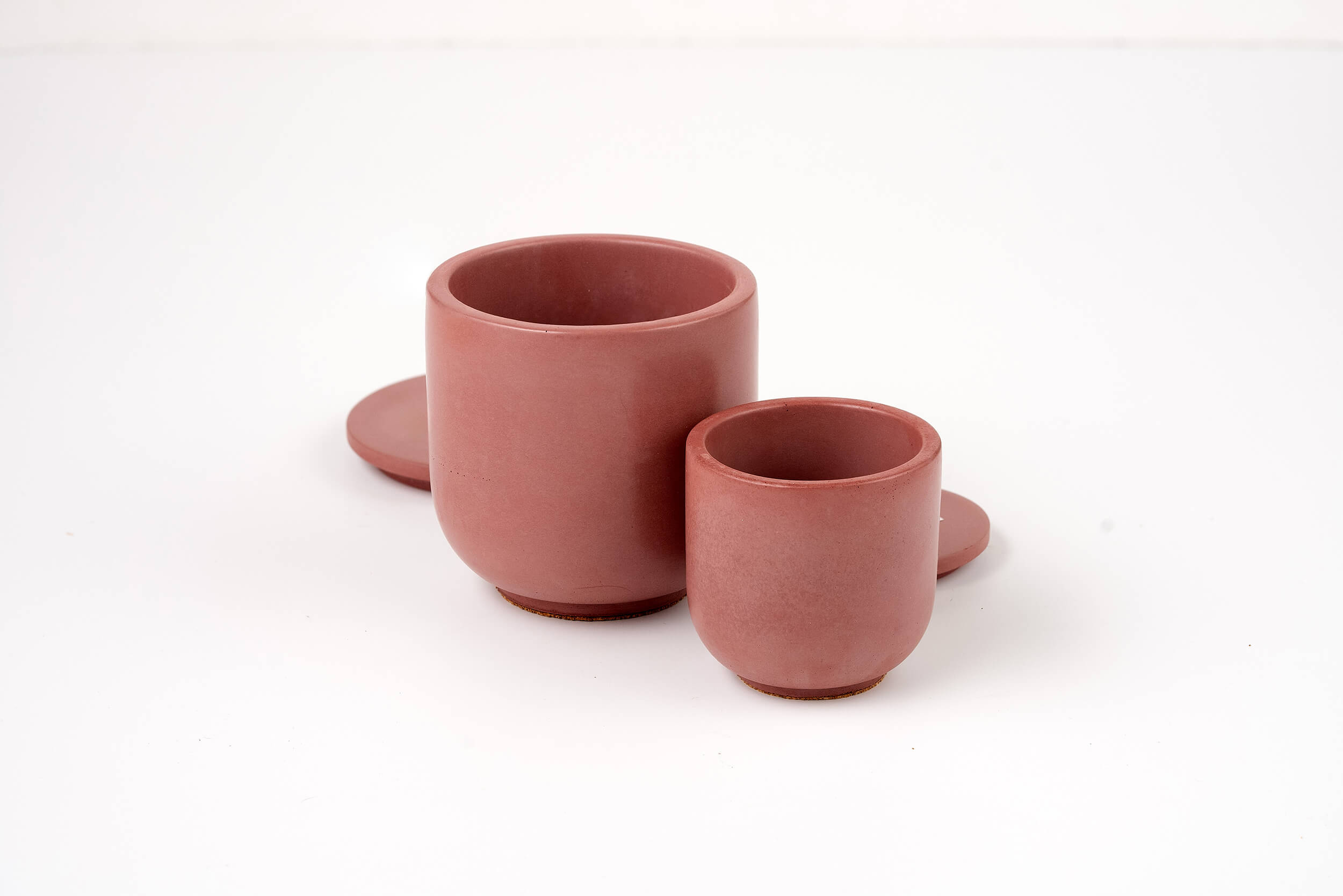 Two minimalist, matte pink ceramic cups of different sizes rest on a white surface, their elegance reminiscent of the Antique Rose Candle Vessel, with matching saucers partially visible behind them.