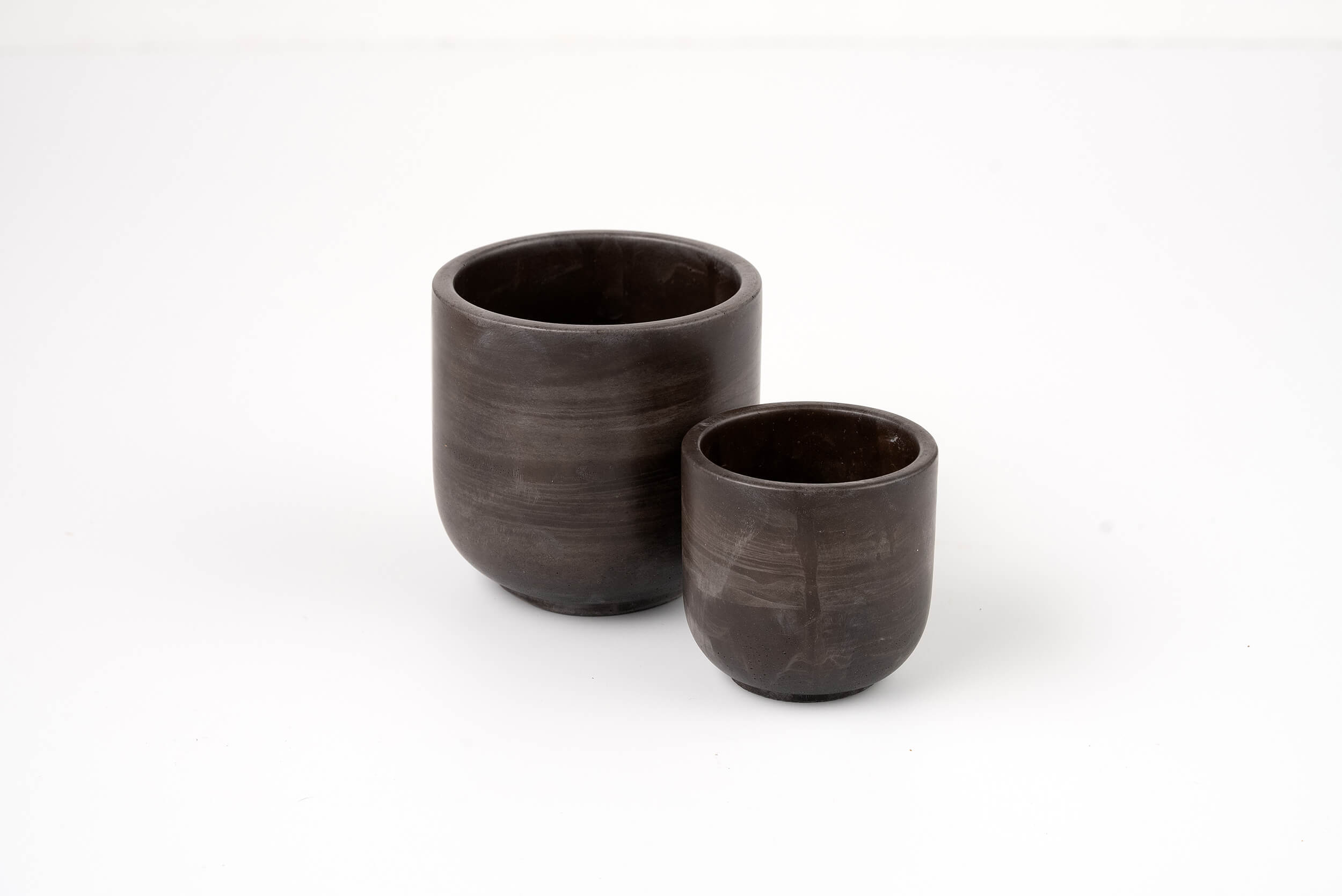 Two dark brown ceramic cups, inspired by the Graphite Candle Vessel, are placed side by side on a plain white background, showcasing minimalist design in different sizes.